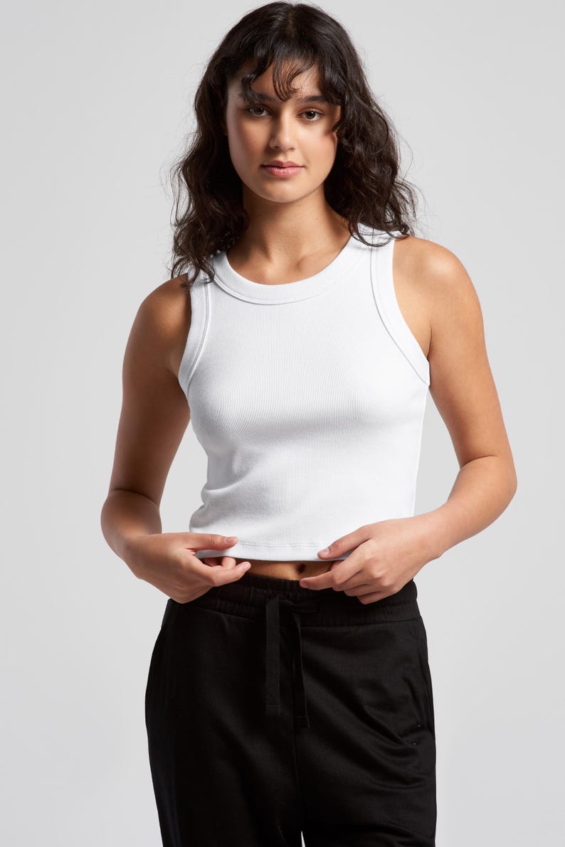 May include: A white ribbed tank top with a crew neck. The top is sleeveless and cropped, and the model is wearing black sweatpants. The top is made of a soft, ribbed material.