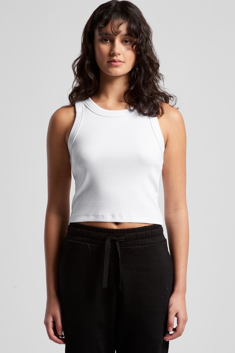May include: A white ribbed tank top with a crew neck. The top is paired with black sweatpants with a drawstring. The model is standing in front of a white background.