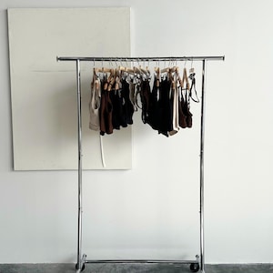 May include: A chrome clothing rack on wheels displays various tops and bottoms in neutral colors. The garments hang on wooden hangers against a white wall. The text "PRICE INCLUDES DUTIES FOR U.S. ORDERS" is visible.