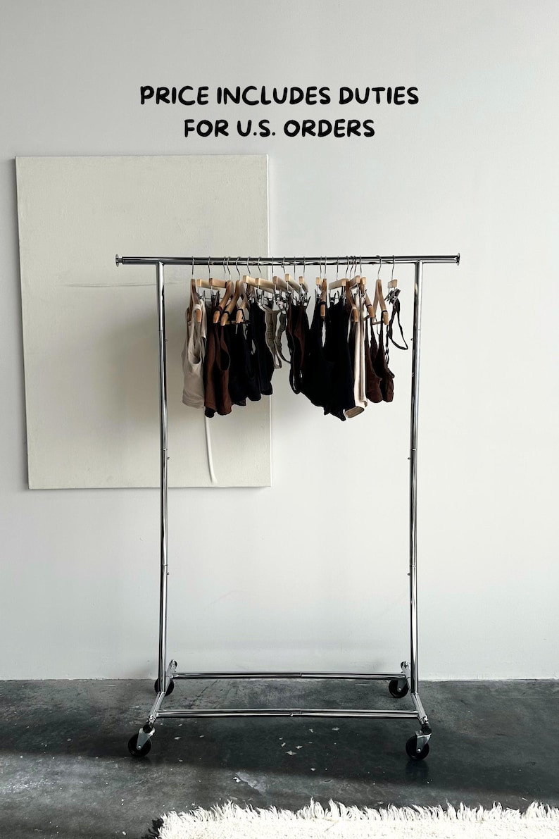 May include: A chrome clothing rack on wheels displays various tops and bottoms in neutral colors. The garments hang on wooden hangers against a white wall. The text "PRICE INCLUDES DUTIES FOR U.S. ORDERS" is visible.
