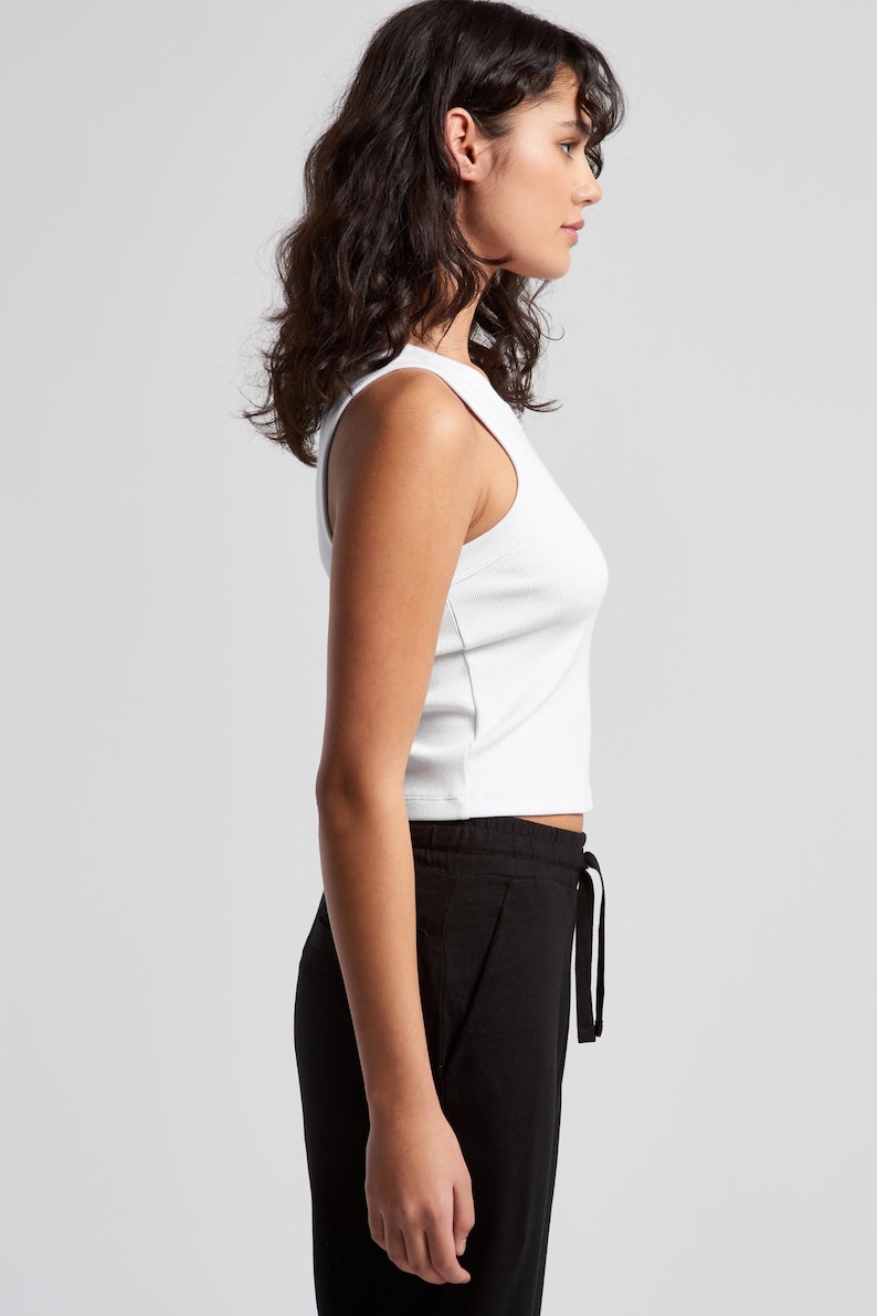 May include: A white ribbed tank top and black drawstring pants are shown in a side view. The tank top is sleeveless and cropped. The pants are loose-fitting with a drawstring waist. The background is plain.