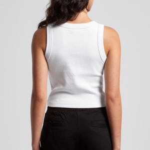 May include: A white ribbed tank top with a scoop neck and wide straps. The tank top is paired with black pants. The model is viewed from the back, with dark hair and a neutral background.