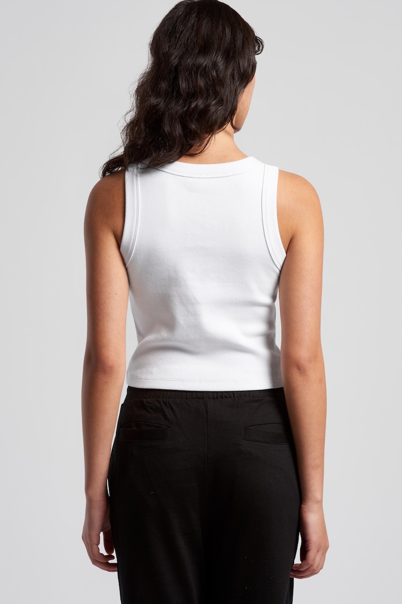 May include: A white ribbed tank top with a scoop neck and wide straps. The tank top is paired with black pants. The model is viewed from the back, with dark hair and a neutral background.
