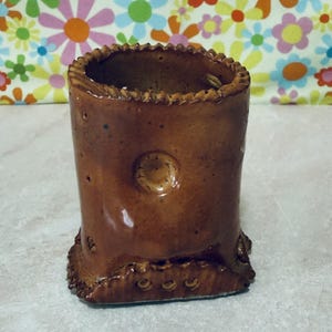 May include: A handmade, cylindrical brown ceramic vase with a textured surface. The vase features a decorative, ridged rim and a circular embossed design on the front. The base has a detailed, scalloped edge. The vase is approximately 5 inches tall.
