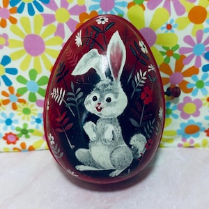 May include: Vintage red Easter egg-shaped tin with a painted white rabbit design. The rabbit has pink-lined ears and a smiling face, surrounded by floral and foliage details. A small, dark knob is on the side.