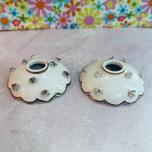 May include: Two white ceramic candle holders with a scalloped edge and gold trim. Each holder has a central opening and is decorated with small, textured floral accents. The background is a colorful floral pattern.