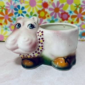 May include: A ceramic hippo planter with a pink and white body, blue eyes, and a decorative collar. The planter has brown feet and a green base. The background features a floral pattern.