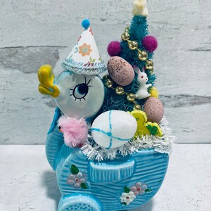 May include: A light blue ceramic duck figurine in a wagon, decorated for Easter. The duck wears a party hat and has a pink chick attached. The wagon holds a small teal tree with Easter eggs, a bunny, and gold beads.