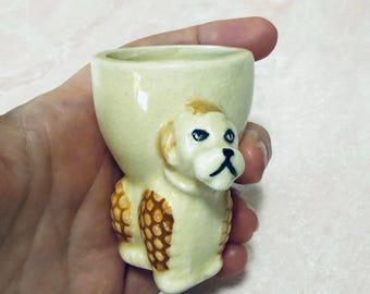 Könnte beinhalten: Ein cremefarbener Eierbecher aus Keramik mit einem Pudel-Design. Der Pudel ist hellbeige mit braunen Akzenten auf Rücken und Ohren sowie schwarzen Details für Augen und Nase. Der Eierbecher wird in einer Hand gehalten.