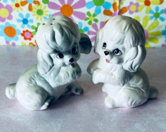 May include: A pair of white ceramic poodle salt and pepper shakers. The shakers have black eyes and pink noses. They are sitting on a floral background.