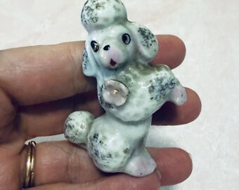 Könnte beinhalten: Kleine Pudelfigur aus Keramik. Der Pudel ist weiß mit grau-schwarzen gesprenkelten Markierungen. Er hat eine rosa Nase und ein Blumendetail auf der Brust. Die Figur wird in einer Hand gehalten.