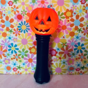 May include: A bright orange plastic jack-o'-lantern head with a carved face featuring triangular eyes and a jagged mouth, mounted on a tall, smooth, cylindrical black handle. The pumpkin head glows with an internal light source.