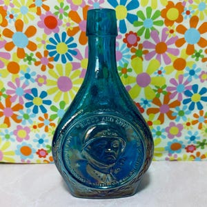 May include: A vintage blue glass bottle with a detailed embossed design. The bottle features a portrait of a man in a military helmet and the words "Guts and Glory". The bottle has a unique shape and a shiny, iridescent finish.