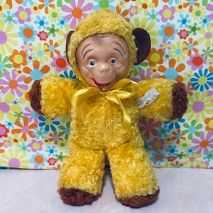 May include: A vintage plush monkey doll with a yellow fur body and a monkey face. The doll has brown ears, hands, and feet, and a yellow ribbon tied around its neck. The doll is set against a colorful floral background.