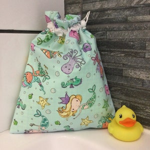 May include: A light green drawstring bag with a mermaid and sea creature print. The bag is made of fabric and has a white drawstring. A yellow rubber duck is sitting on a white surface next to the bag.