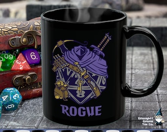 May include: Black ceramic mug with a white design of a Dungeons and Dragons rogue character with a purple cloak and a dagger. The mug has the text "ROGUE" printed on it.