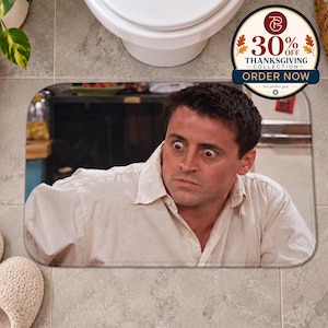 May include: A rectangular bath mat featuring a printed image of a person with wide, surprised eyes wearing a light-coloured button-down shirt. The person is looking towards the left side of the frame with a startled expression on their face.