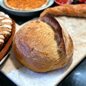 K&ouml;nnte beinhalten: Ein frisch gebackenes Sauerteigbrot mit einer goldbraunen Kruste. Das Brot hat eine leicht unregelm&auml;&szlig;ige Form und einen tiefen Riss auf der Oberseite.