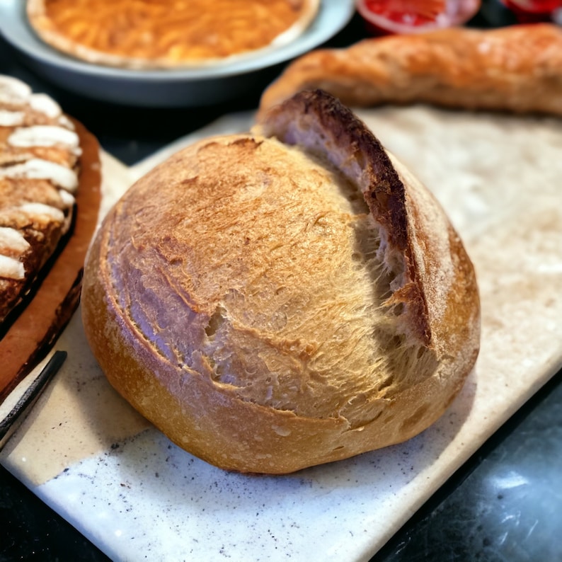 May include: A freshly baked loaf of sourdough bread with a golden brown crust. The bread has a slightly irregular shape and a deep crack on the top.