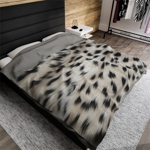 May include: A soft, plush throw blanket with a leopard print pattern in shades of beige and black. The blanket is draped over a bed with a gray headboard.