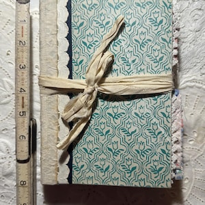 Op de afbeelding: Een handgemaakt dagboek met een teal en witte bloemenprint op de omslag. Het dagboek is vastgebonden met een crèmekleurig lint en heeft een kantversiering.