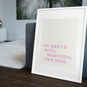 May include: White framed art print with the text "WE SHOULD HANG SOMETHING COOL HERE" in pink. The artwork is leaning against a gray couch on a dark wood floor. A white rug and a silver vase are visible in the background.
