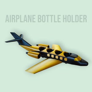 Puede incluir: Soporte para botellas con forma de avión en oro y negro. El soporte para botellas tiene alas, cola y cabina. El texto "AIRPLANE BOTTLE HOLDER" está encima del avión.