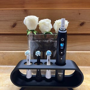 May include: A black matte toothbrush stand with four circular slots holding three electric toothbrush heads and one black Oral-B iO electric toothbrush handle with a digital display showing a blue tooth icon and the text Limpeza Diaria.