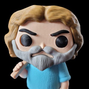 May include: A Funko Pop figure of a man with a light brown beard and hair, wearing a blue sweater and pants. The figure has black eyes and a neutral expression. The figure is holding a light brown object.