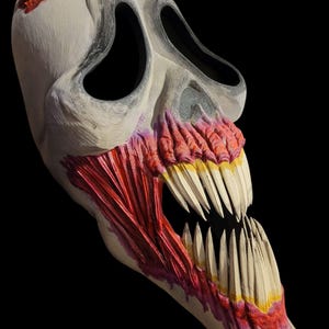 Peut inclure: Un masque de crâne blanc cassé détaillé avec de grandes orbites et une large bouche ouverte. Le masque présente des dents pointues et des tissus musculaires rouges exposés. Le haut du crâne comporte trois cornes pointues à bout jaune. Le masque est sur fond noir.