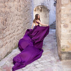 Może przedstawiać: Kobieta w powłóczystej, głębokofioletowej sukni idzie kamienną alejką. Suknia ma odkryte plecy i długi tren, który ciągnie się za nią. Alejka jest wąska, z kamiennymi ścianami po obu stronach i łukowatym przejściem w oddali.