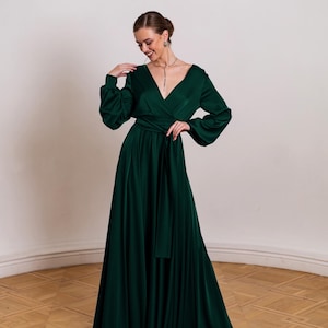 May include: A floor-length emerald green dress with a deep V-neckline, long voluminous sleeves with fitted cuffs, and a wrap-style bodice featuring a matching fabric tie belt at the waist. The skirt flows into a full, floor-length hemline.