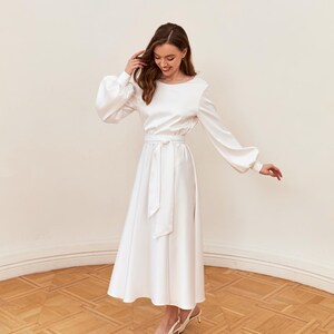 May include: A white satin dress with long sleeves and a midi-length skirt. The dress features a round neckline, a tie-waist belt, and a slight flare at the hem. The model is wearing white heels. The dress is suitable for special occasions.