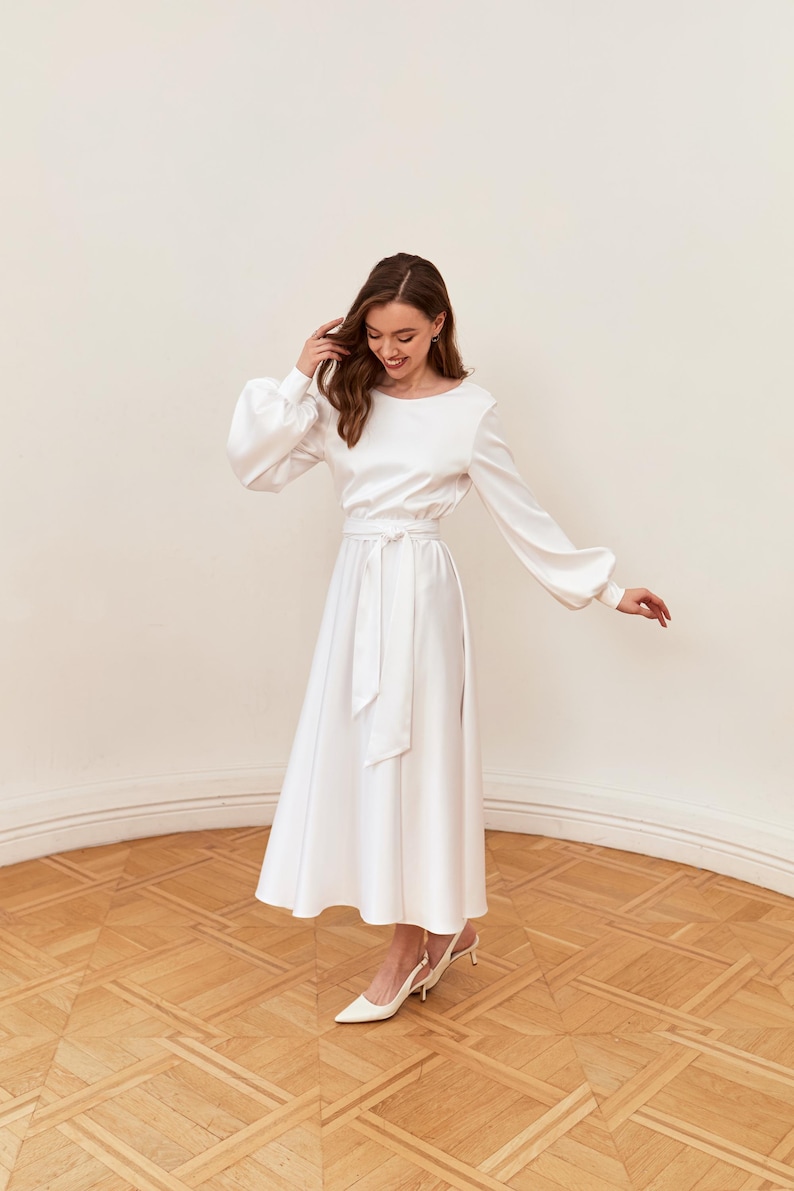 May include: A white satin dress with long sleeves and a midi-length skirt. The dress features a round neckline, a tie-waist belt, and a slight flare at the hem. The model is wearing white heels. The dress is suitable for special occasions.