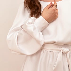 May include: A white dress with long sleeves and a tie waist. The sleeves have a unique, voluminous design. The dress is made of a smooth, flowing fabric. The overall style is elegant and minimalist.