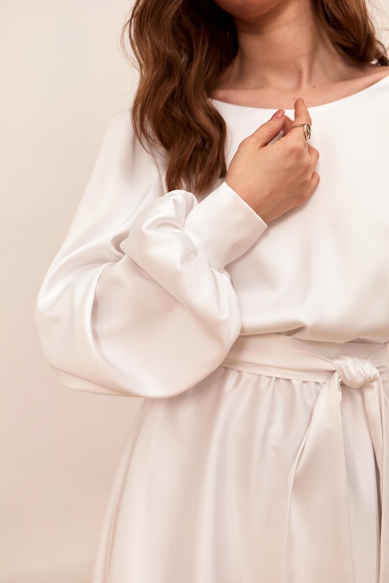 May include: A white dress with long sleeves and a tie waist. The sleeves have a unique, voluminous design. The dress is made of a smooth, flowing fabric. The overall style is elegant and minimalist.