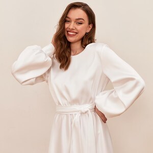 May include: A white satin dress with long sleeves and a tie waist. The dress features a round neckline and voluminous sleeves. The model is smiling and has her hand on her head. The dress is a midi length.