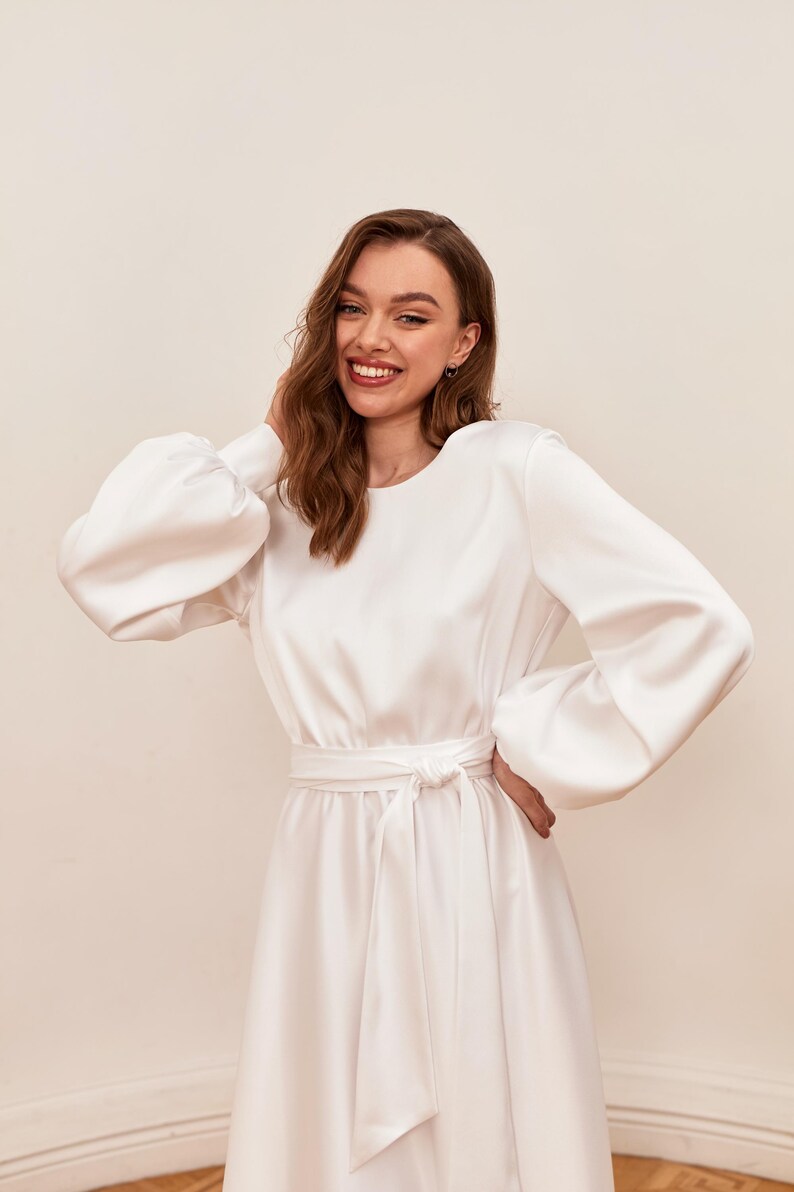May include: A white satin dress with long sleeves and a tie waist. The dress features a round neckline and voluminous sleeves. The model is smiling and has her hand on her head. The dress is a midi length.