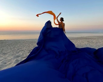 May include: A person standing on a beach with a blue fabric draped over them. The fabric is flowing in the wind, and the person is holding a long orange scarf in their hand.