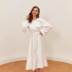 May include: A white satin dress with long, puffy sleeves and a midi-length skirt. The dress features a tie-waist and is paired with white pointed-toe heels. The dress is suitable for formal events.