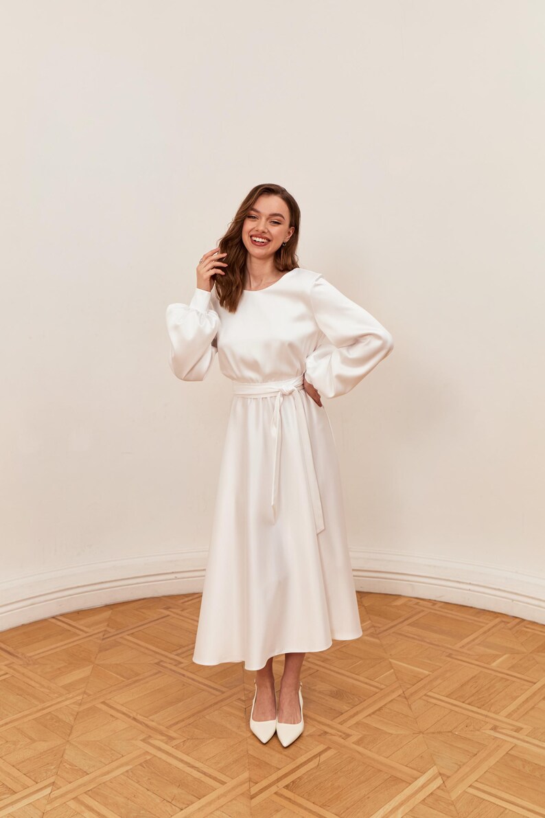 May include: A white satin dress with long, puffy sleeves and a midi-length skirt. The dress features a tie-waist and is paired with white pointed-toe heels. The dress is suitable for formal events.