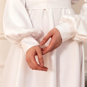 Może przedstawiać: Biała sukienka z długimi rękawami i dopasowaną talią. Rękawy mają bufiasty, marszczony wygląd. Sukienka ma sp&oacute;dnicę w stylu falban.