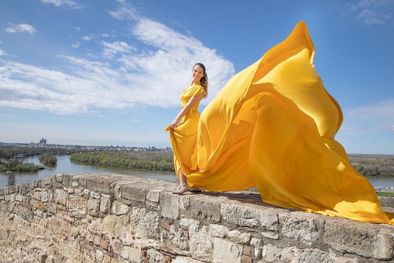 May include: A woman in a bright yellow dress stands on a stone wall overlooking a river and city. The wind is blowing her dress, creating a dramatic effect.