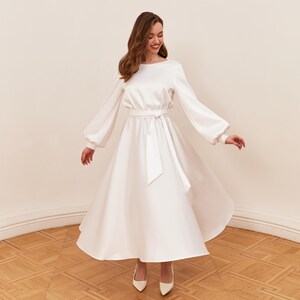 May include: A white satin dress with long sleeves and a flowing skirt. The dress features a boat neckline, a cinched waist with a matching belt, and a midi-length hemline. The sleeves have a slight puff at the shoulders and gather at the wrists. The model is wearing white heels.