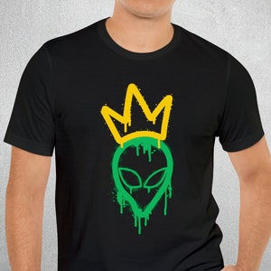 Puede incluir: Camiseta negra con un gráfico de cabeza de extraterrestre verde con una corona amarilla. La cabeza del extraterrestre gotea pintura verde.