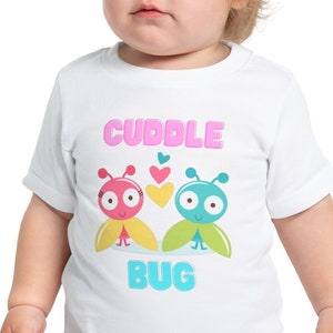 May include: White toddler t-shirt with a pink, yellow, and blue graphic of two cartoon bugs with hearts above them. The text "Cuddle Bug" is printed in pink letters on the shirt.