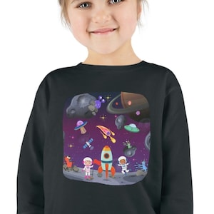 Puede incluir: Una sudadera negra de manga larga con un estampado gráfico colorido de astronautas, cohetes y planetas en el espacio. El gráfico está centrado en la parte delantera de la sudadera.