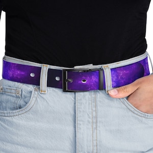 Peut inclure: Une ceinture à imprimé galaxie violet et noir avec une boucle argentée. La ceinture est portée sur un jean bleu clair.