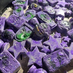 May include: A collection of purple and green wax melts with various Halloween-themed shapes, including stars, coffins, and a pumpkin. The wax melts have a glittery finish.
