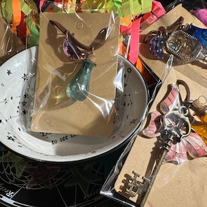 May include: Assortment of handmade glass bottle charms on necklaces and keychains. The charms are filled with colourful liquids and glitter. The necklaces and keychains are displayed on brown cardstock with white text.
